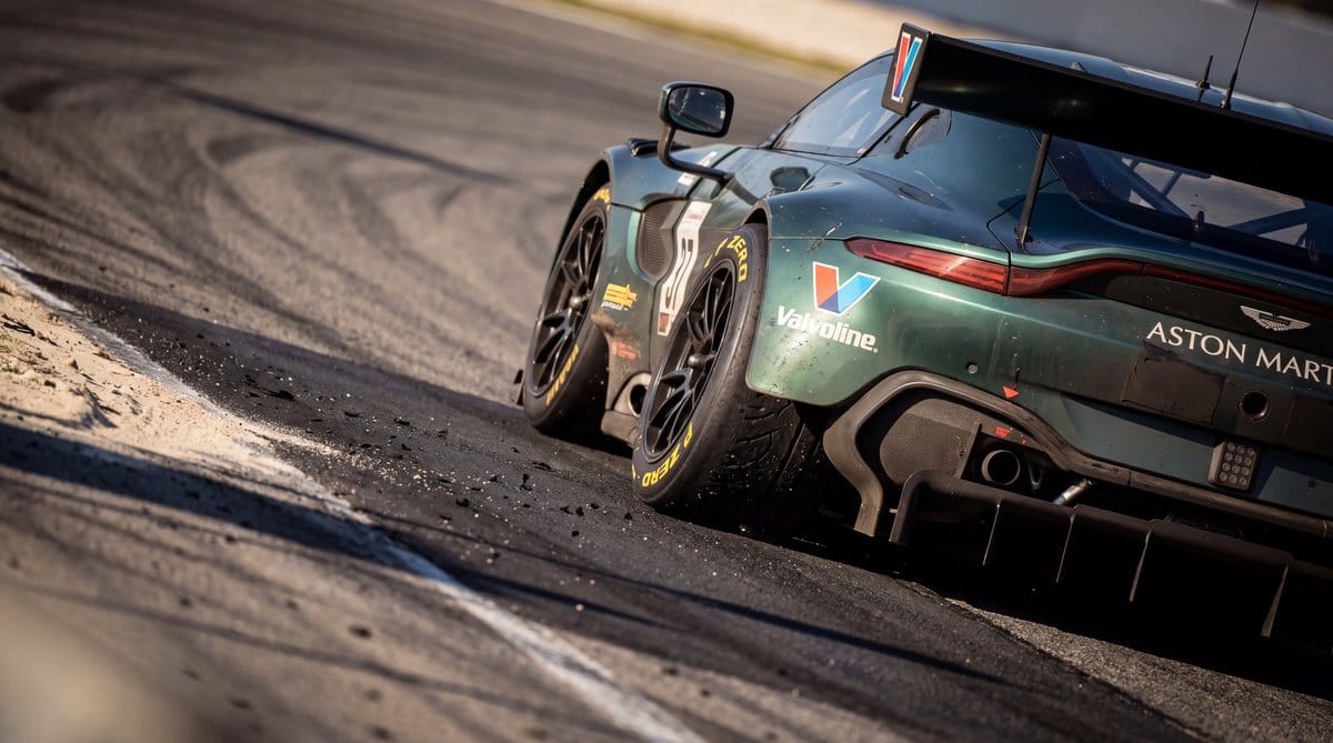 Aston Martin F1 car’s rear tyres deform on Turn 3’s steep banking beside scattered beach sand.
