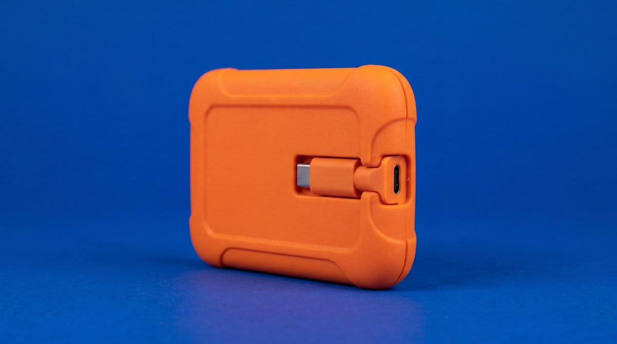 An orange rugged portable solid-state drive with an integrated connector stands against a deep cobalt background.