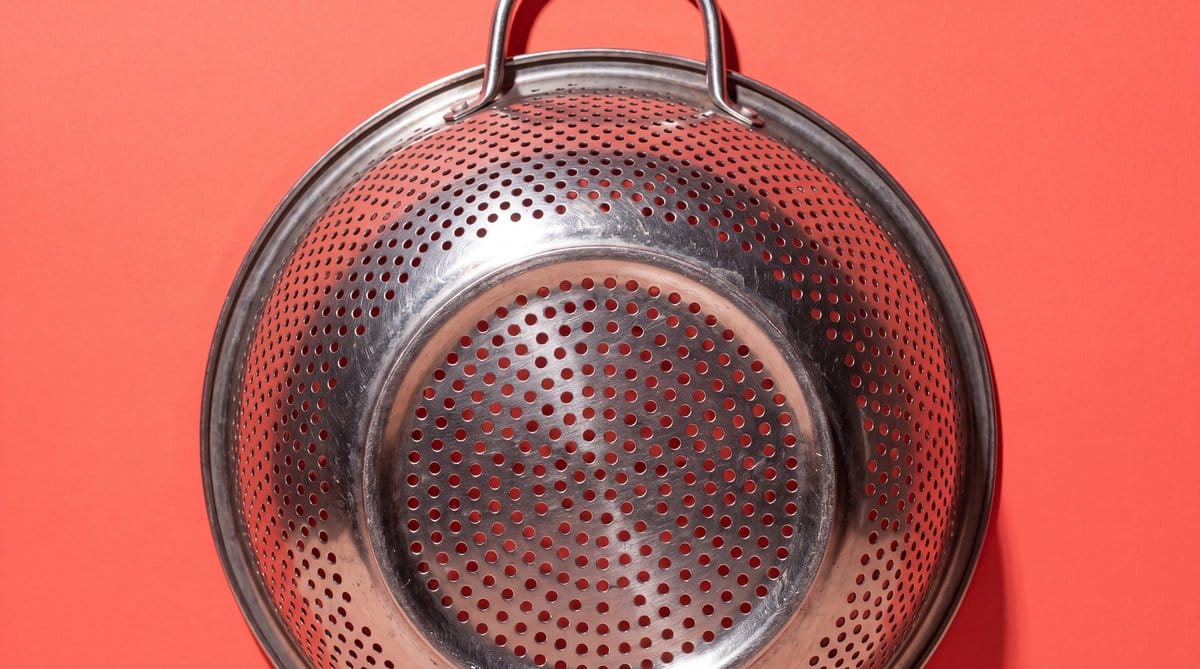 Stainless-steel colander standing upright against a saturated coral background, its perforated bowl filling the frame.