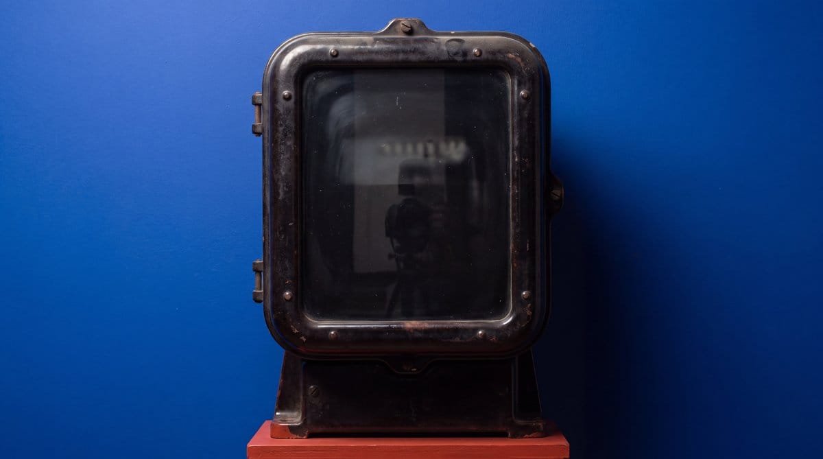 Large dark industrial electricity meter with an obscured glass display, standing on a red base against blue.