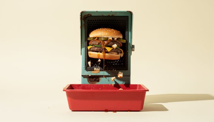 Weathered turquoise drive-thru speaker crushing a burger above a red tray as crumbs and sauce fall.