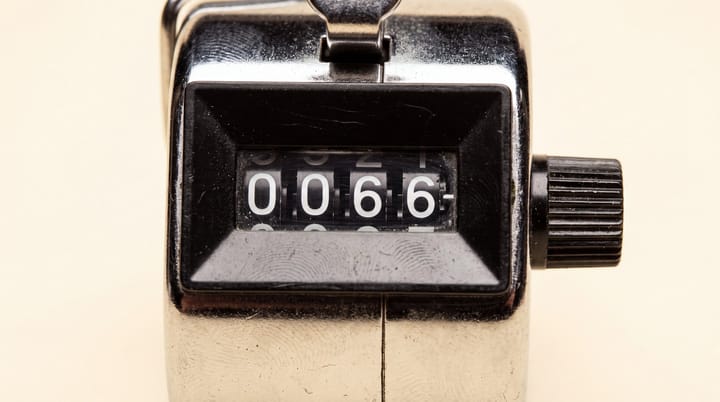 Extreme close-up of a worn chrome mechanical tally counter displaying 0066 against a warm ivory background.